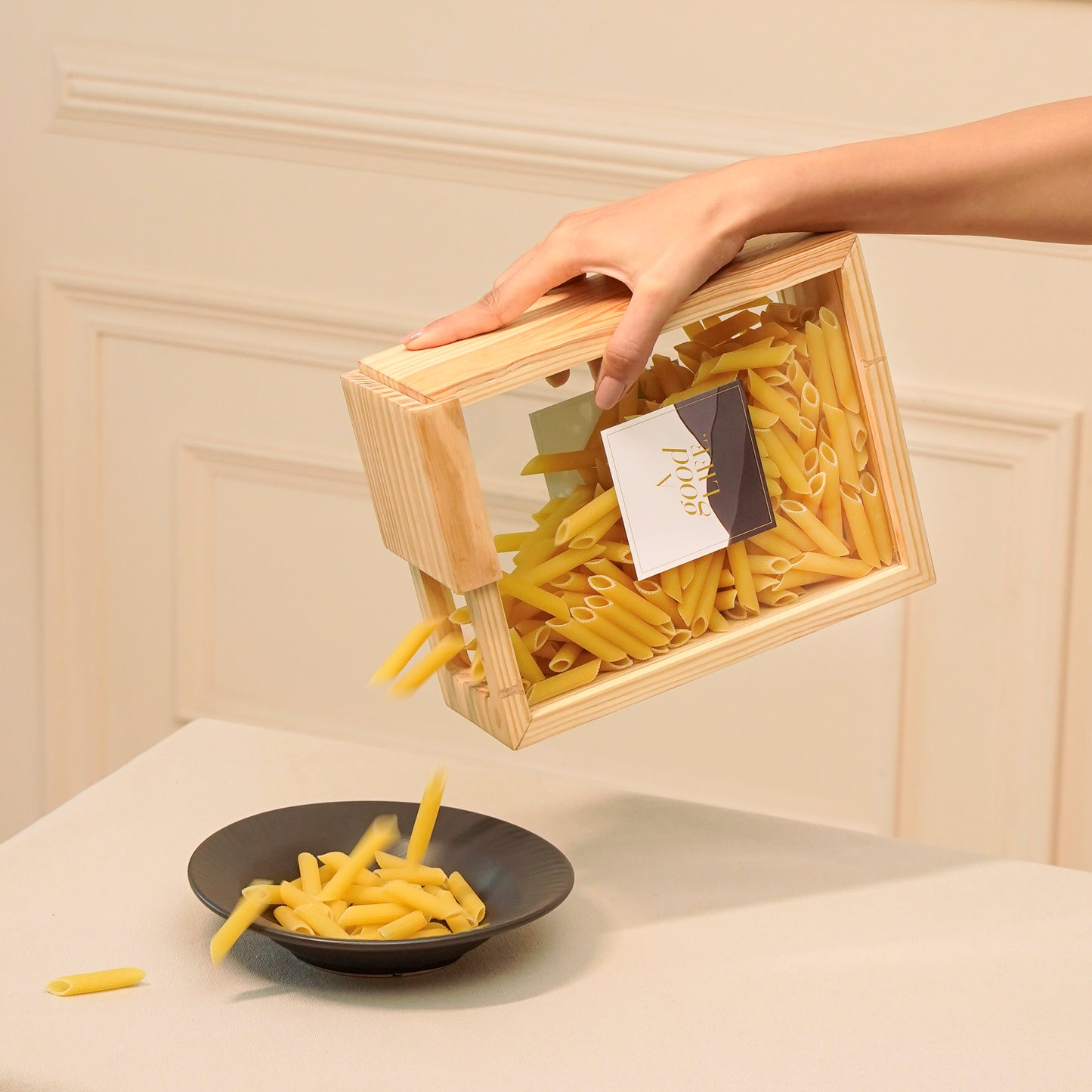 Hand pouring pasta from a pine pantry box by A Good Life into a black bowl on a table