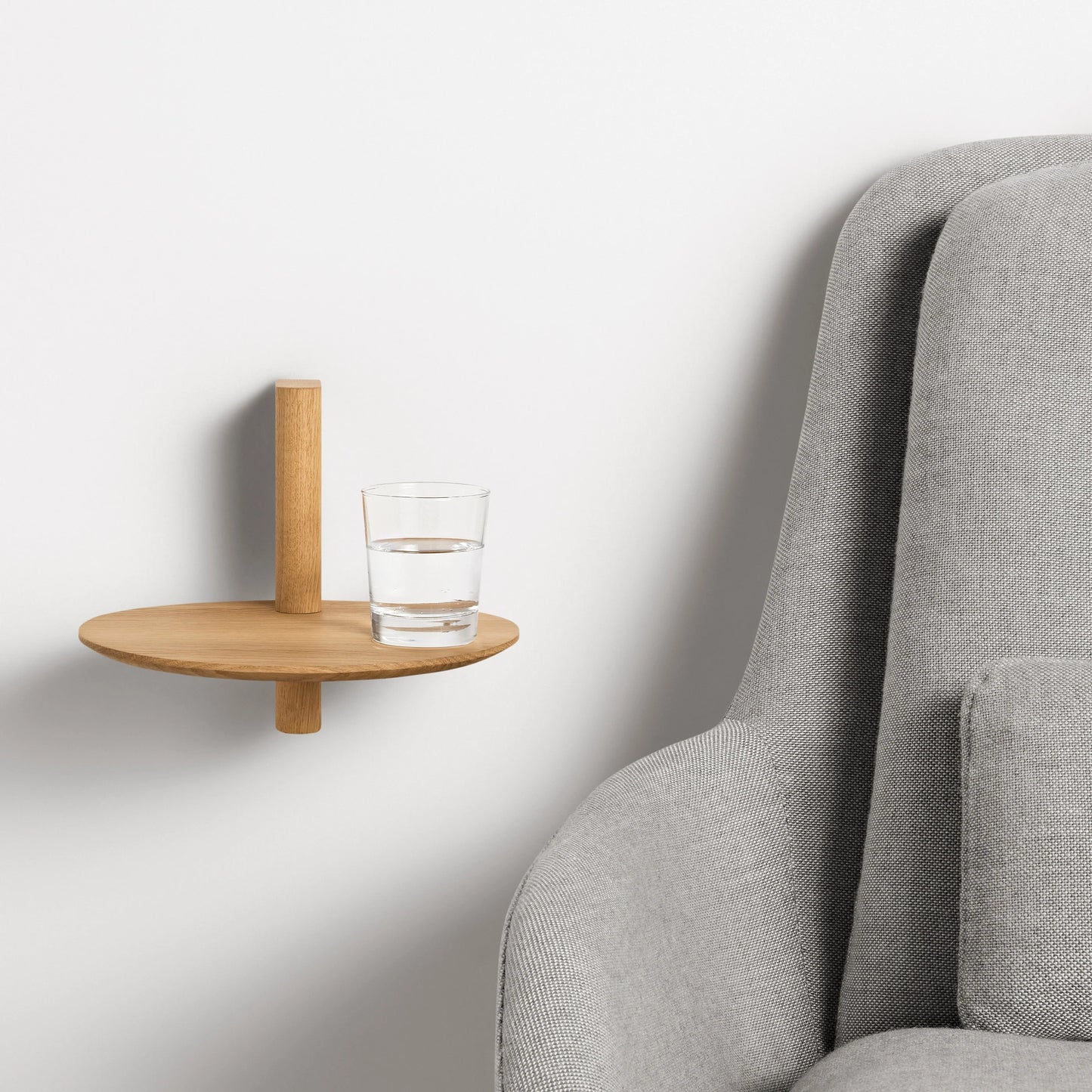 Minimalist pine wood floating shelf mounted on wall holding a glass of water beside a gray upholstered chair