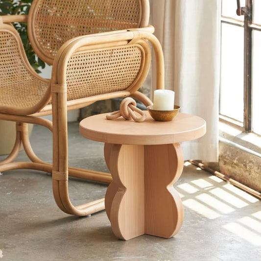 A Good Life minimalist wood nightstand - HUSTLE with a round top and sculpted base beside a wicker chair in natural light