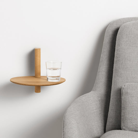 Minimalist pine wood floating shelf mounted on wall holding a glass of water beside a gray upholstered chair